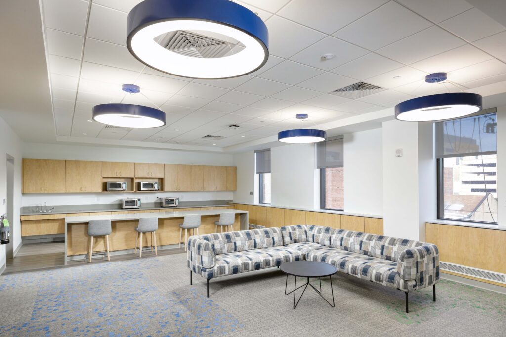 A modern office lounge with a plaid sectional sofa, a small round table, and a kitchen counter with bar stools under circular pendant lights.