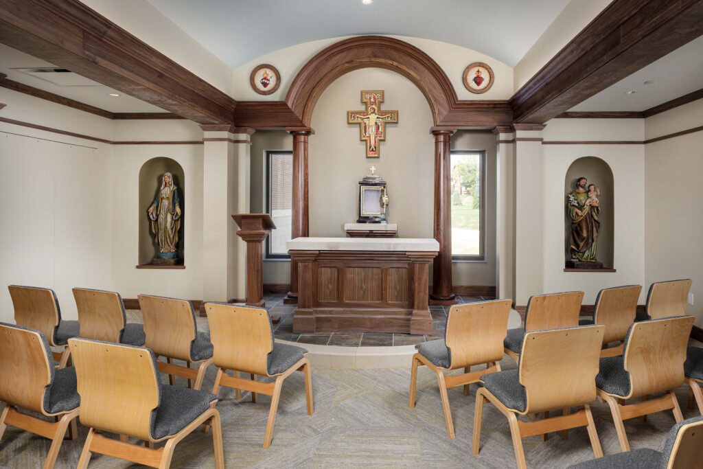 A chapel interior with an altar, a crucifix, and two statues of saints in niches, flanked by rows of wooden chairs.