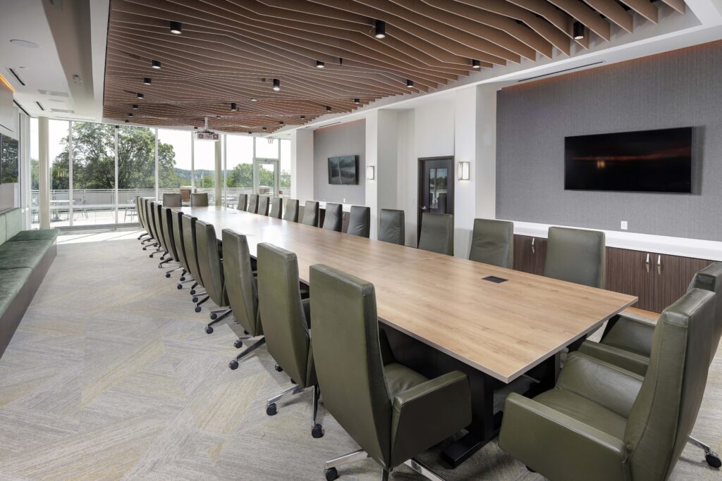 A long wooden conference table surrounded by green leather chairs in a room with a slatted wood ceiling and large windows.