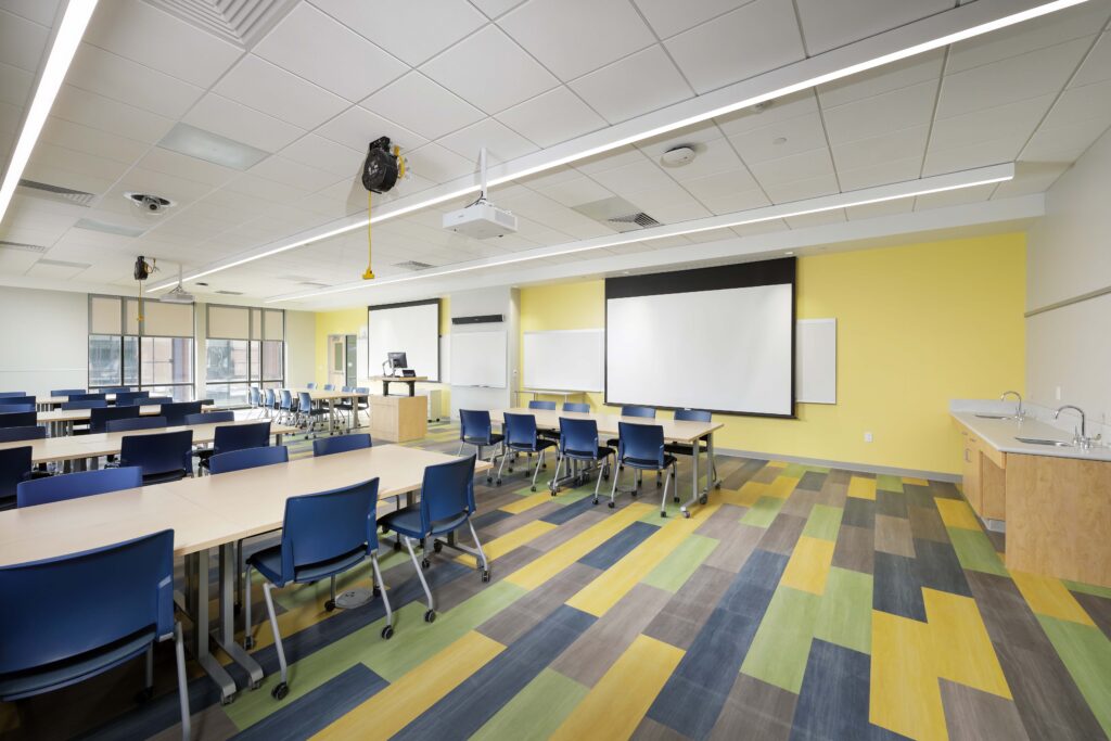 A classroom with rows of tables and blue chairs, a projector screen on a yellow wall, and multicolored floor tiles.