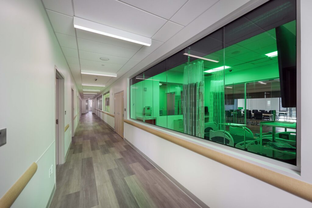 Hospital hallway leading to a glass-walled room illuminated by green light, showing patient beds and medical equipment.