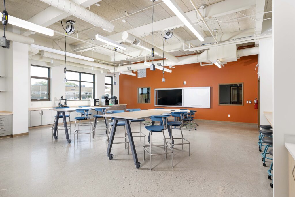 A modern science classroom featuring modular tables with blue stools, an orange accent wall, a whiteboard, and exposed industrial ceiling pipes.