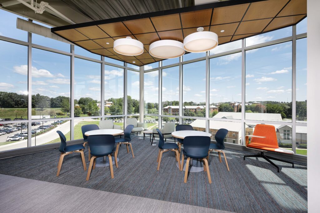 Meeting area with two circular tables, blue chairs, and one orange accent chair, surrounded by floor-to-ceiling windows overlooking a parking lot and campus buildings.
