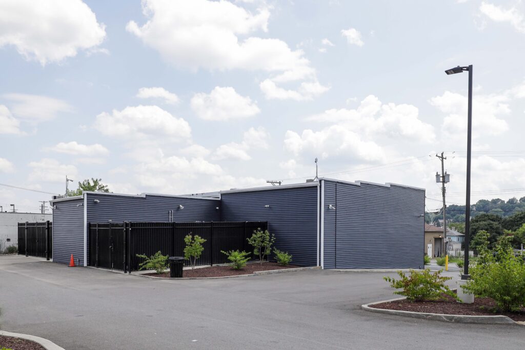 A single-story commercial building with dark blue horizontal siding, a fenced enclosure, and an adjacent asphalt parking lot.