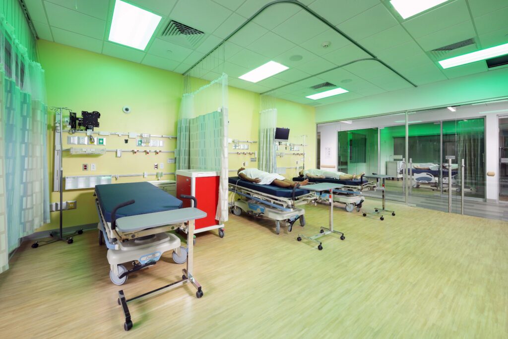 A hospital simulation room with three medical beds, two of which have patient manikins, and wall-mounted medical equipment.
