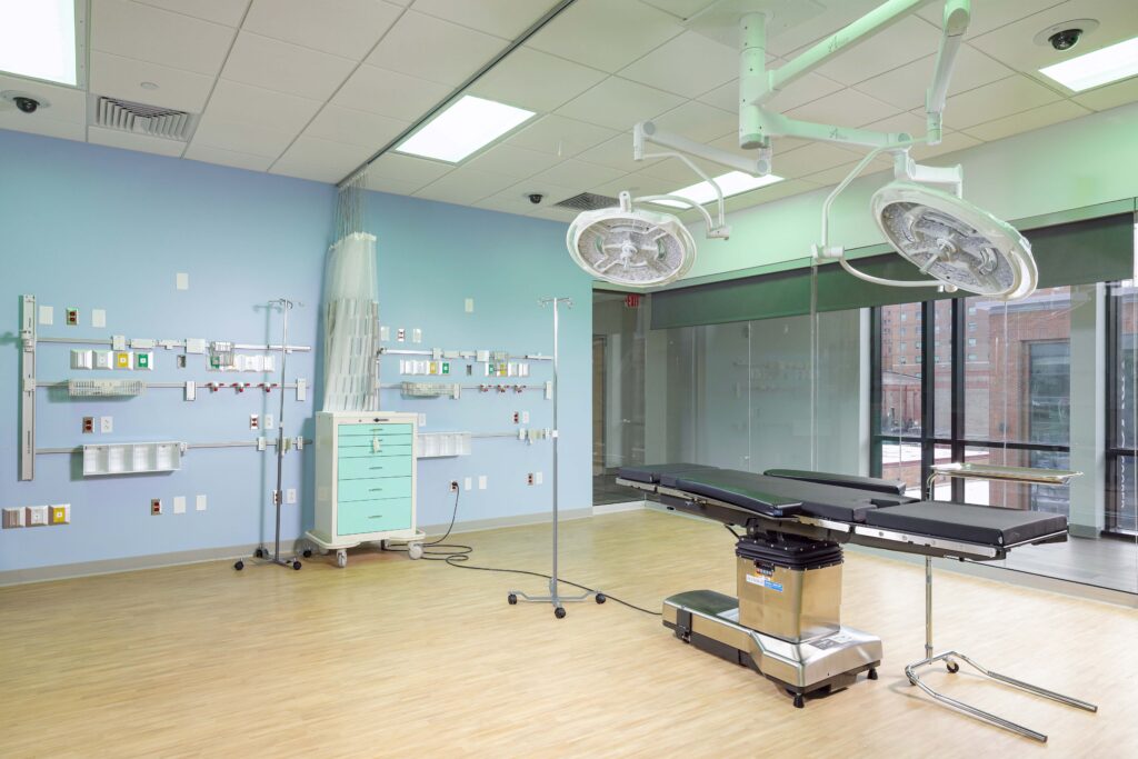 An operating room featuring a surgical table in the center, ceiling-mounted surgical lights, and medical equipment supply units mounted on a light blue wall.