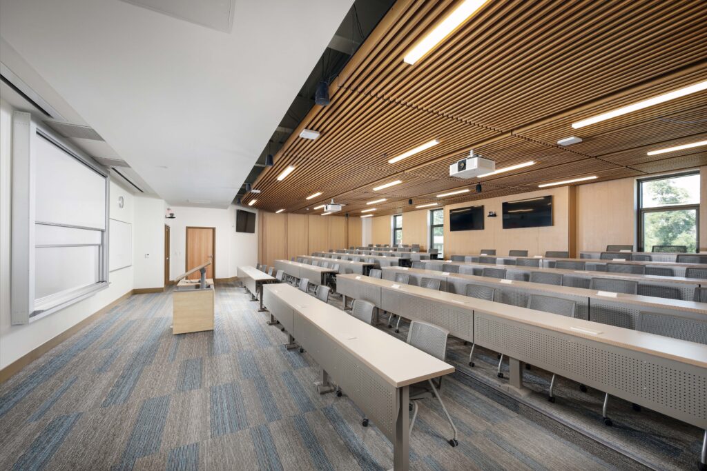 A tiered university lecture hall with rows of desks facing a podium and a large projection screen.