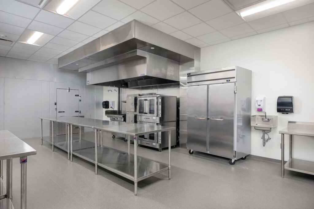 A commercial kitchen featuring stainless steel work tables, a double-stack convection oven under a ventilation hood, and an upright refrigerator.