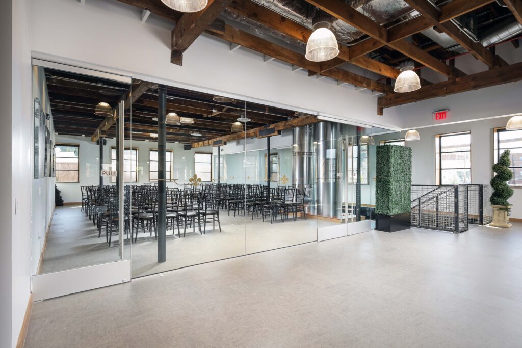 Empty event space viewed through a glass wall, featuring rows of black chairs, exposed ceiling beams, and pendant lighting.