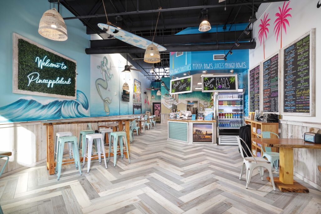 Interior of a tropical-themed cafe with a surfboard hanging from the ceiling, a mermaid mural, and a menu board listing bowls and juices.