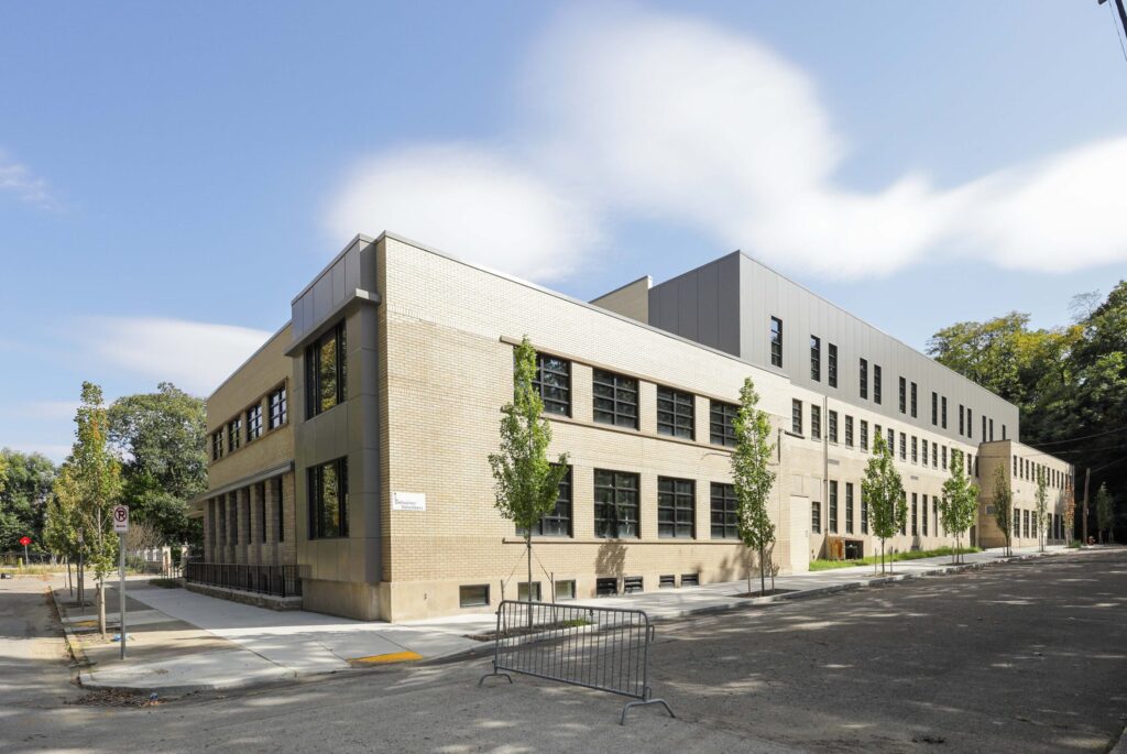 A long, rectangular building with tan brick and grey siding on a street corner, framed by young trees and a metal barricade.