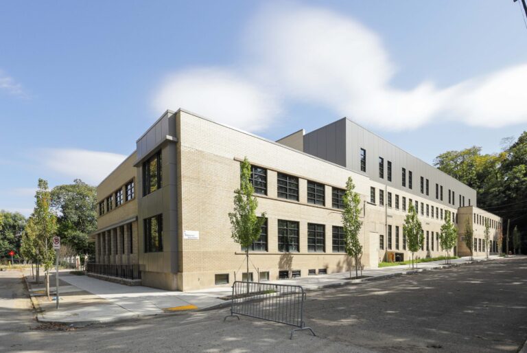 A long, rectangular building with tan brick and grey siding on a street corner, framed by young trees and a metal barricade.