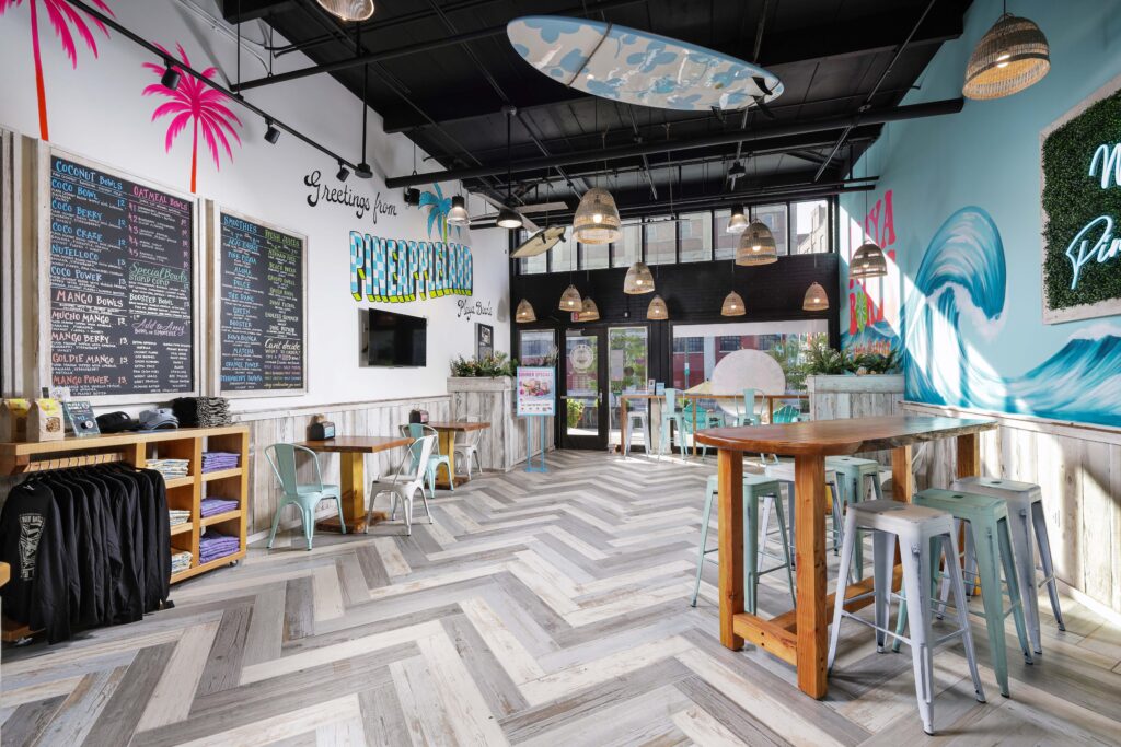 Interior of a tropical-themed cafe with menu boards on the wall, seating, and a surfboard mounted on the ceiling.
