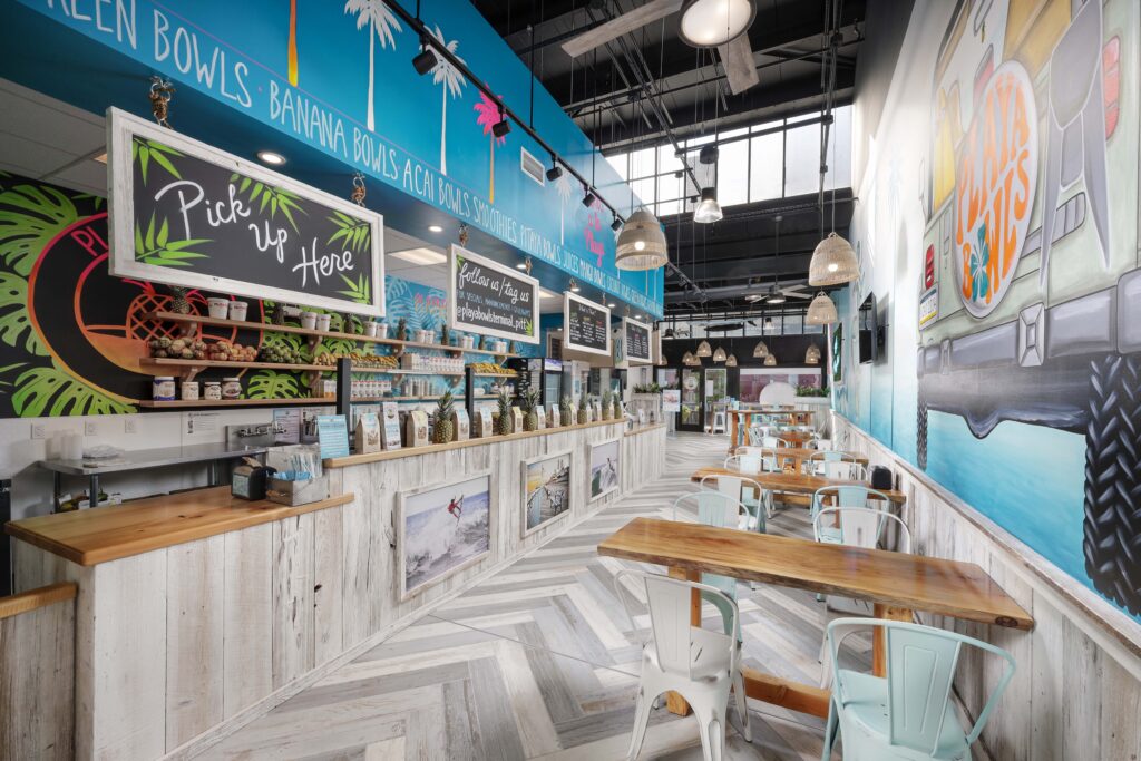 The interior of a Playa Bowls restaurant featuring a wooden service counter with a “Pick up Here” sign, bar stools, and tables along a wall painted with a large mural of a surf van.