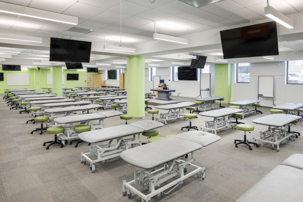 Large classroom or lab featuring rows of adjustable treatment tables with rolling stools, flat-screen monitors mounted on the ceiling, and several whiteboards.