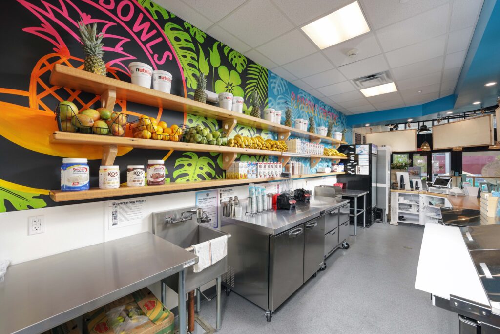 The interior of a juice or smoothie bar featuring stainless steel prep tables, a sink, and long wooden shelves stocked with fresh fruit, Nutella, and protein powder against a colorful wall mural.