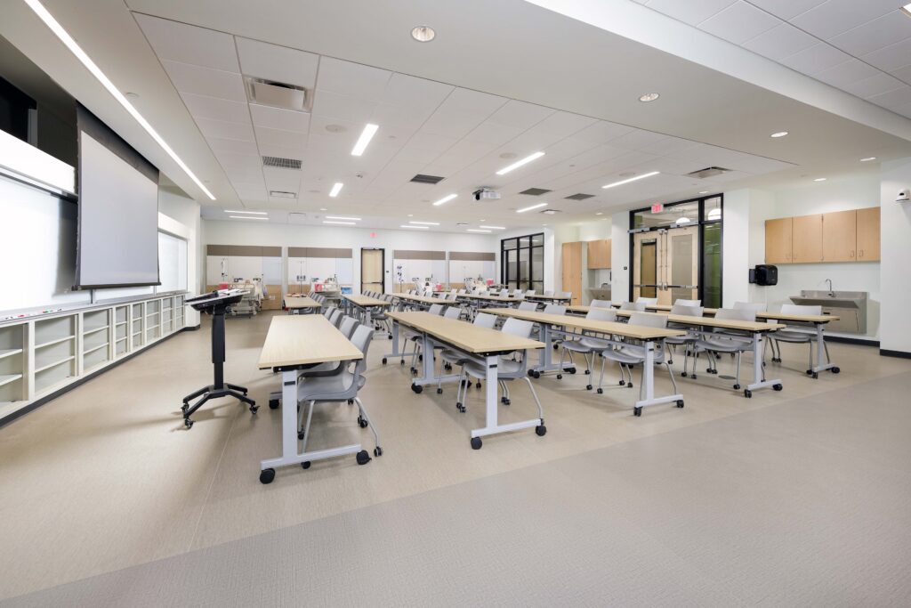 A training classroom with rows of tables, rolling chairs, a projection screen, and medical manikins in the background.