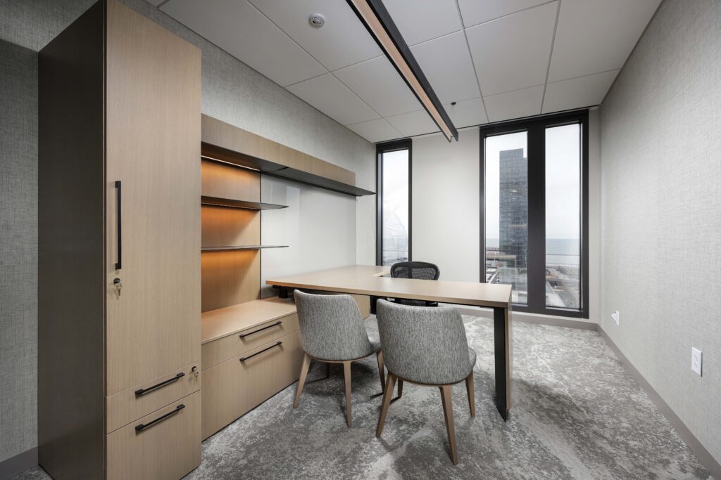 A modern office featuring a wooden desk, two grey upholstered chairs, and integrated storage shelves, with a view of a city skyline through the window.