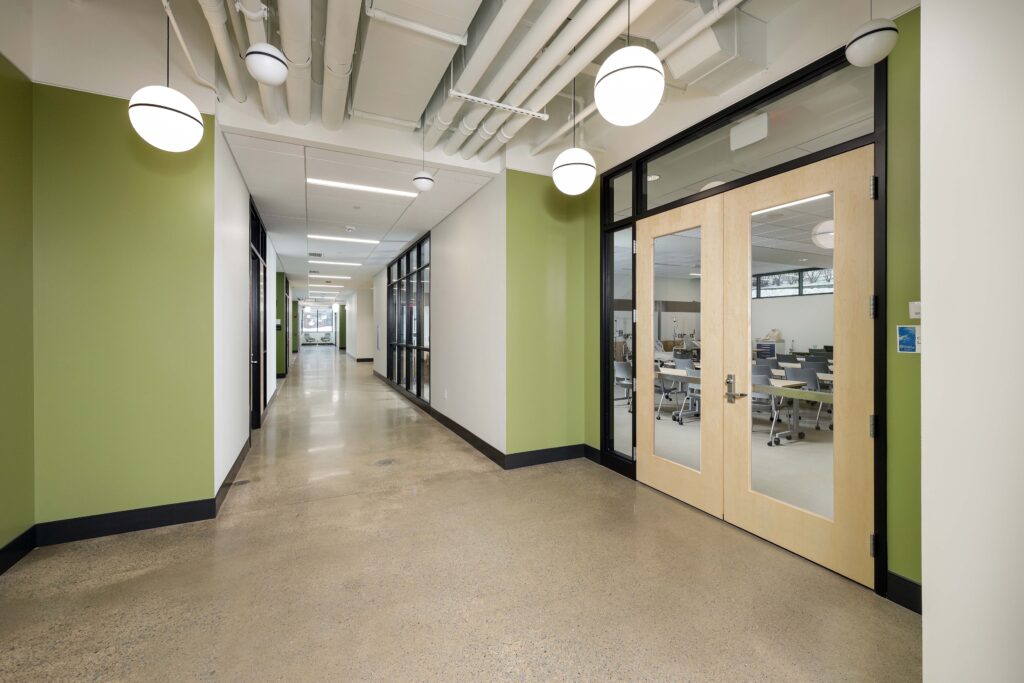 A wide hallway with green walls and light wood double doors opening into a classroom with desks and chairs.