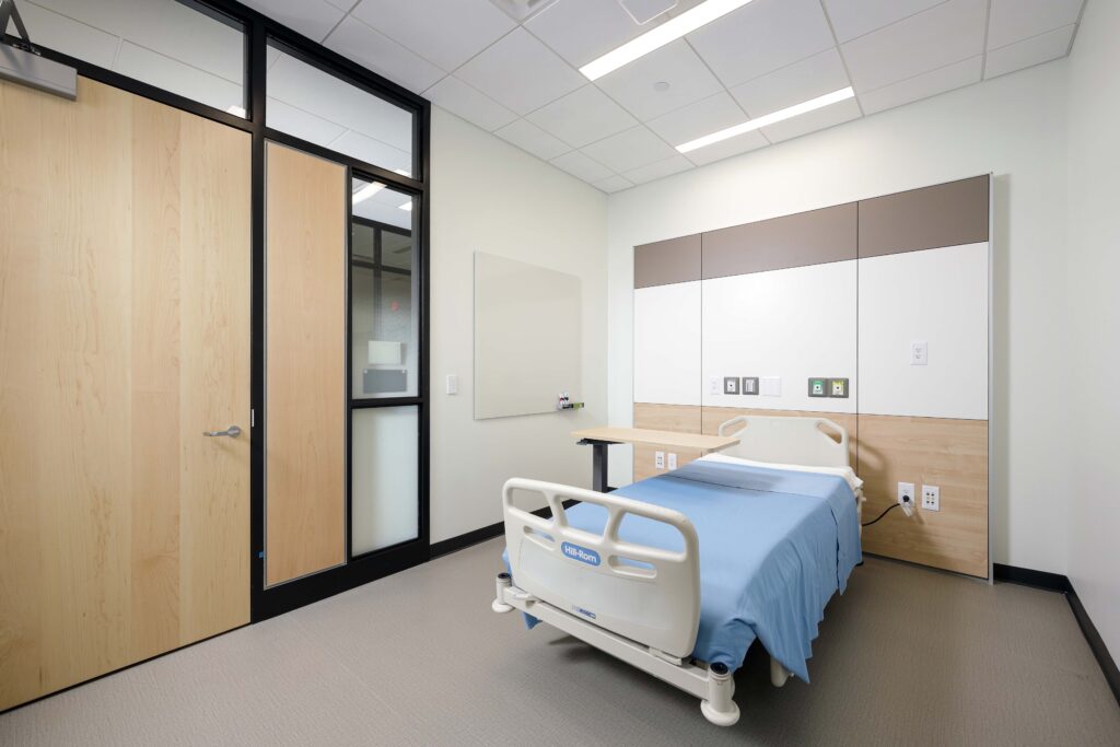 A hospital room with a Hill-Rom patient bed, a wooden door, and medical wall panels with power and equipment outlets.