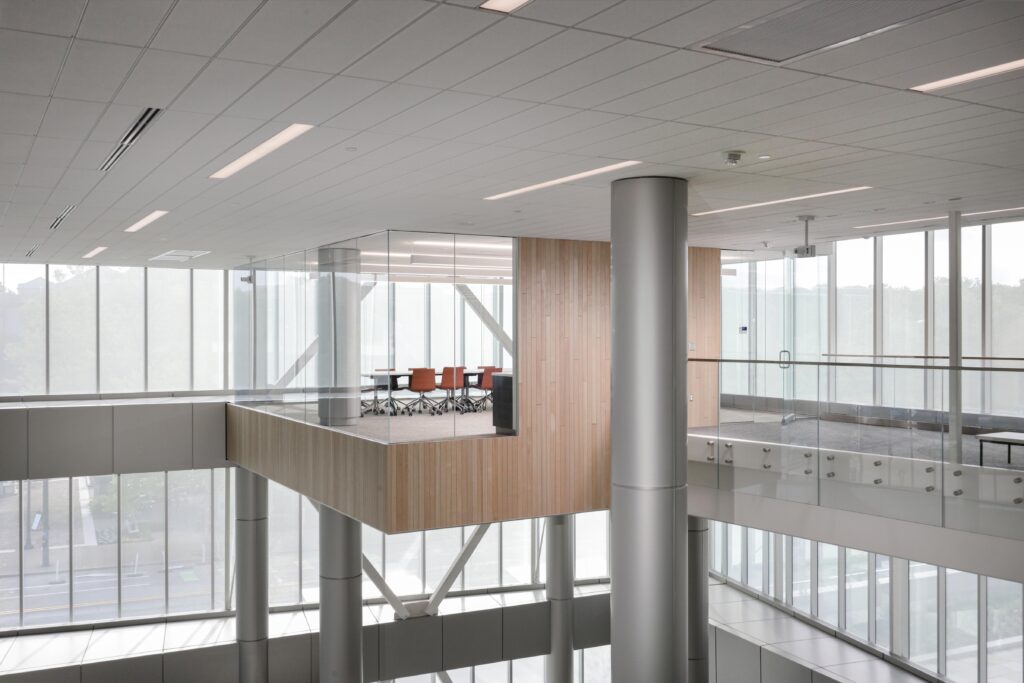A glass-enclosed conference room with orange chairs, cantilevered within a modern office building atrium.
