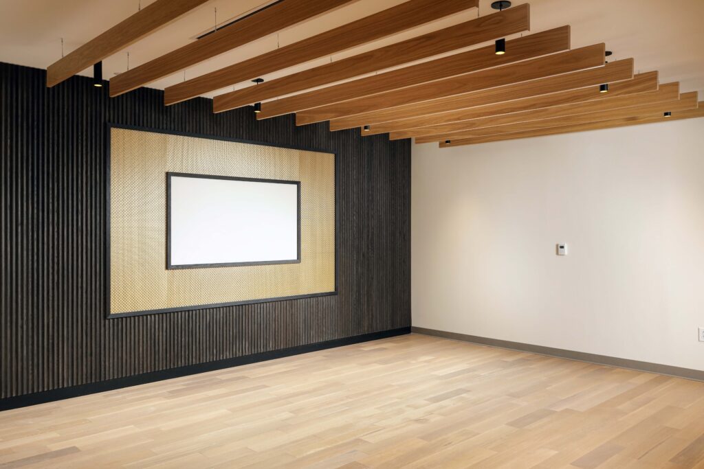 Empty room with light wood floors, dark ribbed walls, and a ceiling featuring wood slats and integrated lighting. A rectangular framed screen is mounted on the wall.