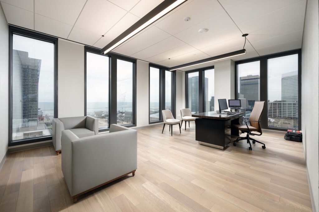 A modern office with a large desk, an ergonomic chair, two guest chairs, and two armchairs, featuring floor-to-ceiling windows with a city view.