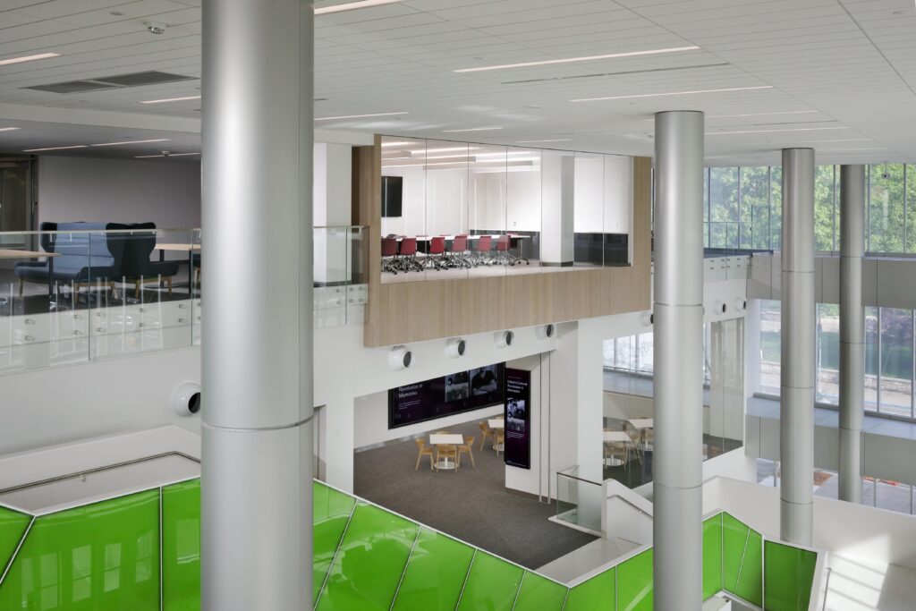 An interior view of a modern building with a multi-level atrium, including a glass-walled conference room, open seating areas, and bright green panels.
