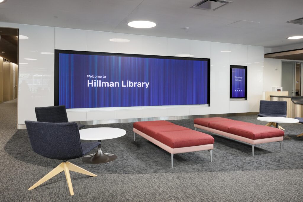 Lobby of Hillman Library featuring two red benches, two chairs with small tables, and digital displays on the wall.
