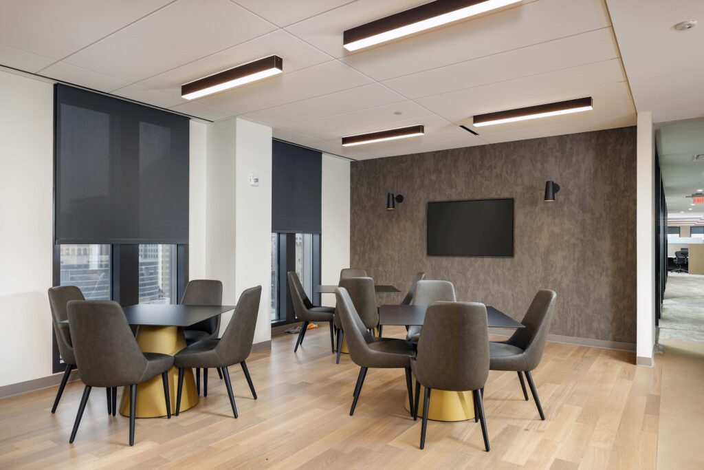 Two tables with grey chairs and gold-tone bases in a modern office break room, with a wall-mounted television.