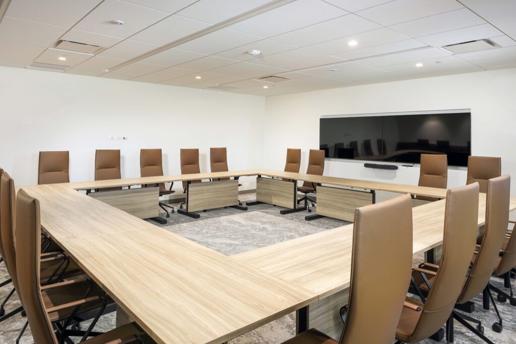 A U-shaped conference table surrounded by brown chairs in a brightly lit office meeting room with a wall-mounted monitor.