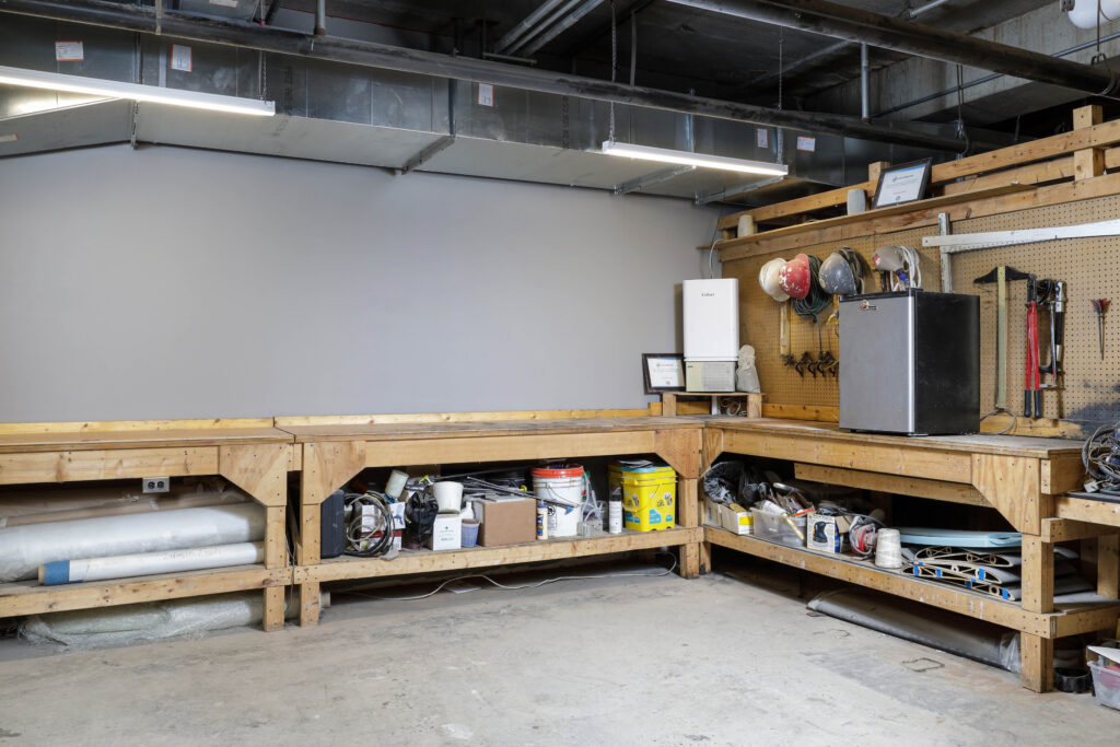 An L-shaped wooden workbench in a workshop with various tools, buckets, and materials stored on the shelves and a pegboard wall.