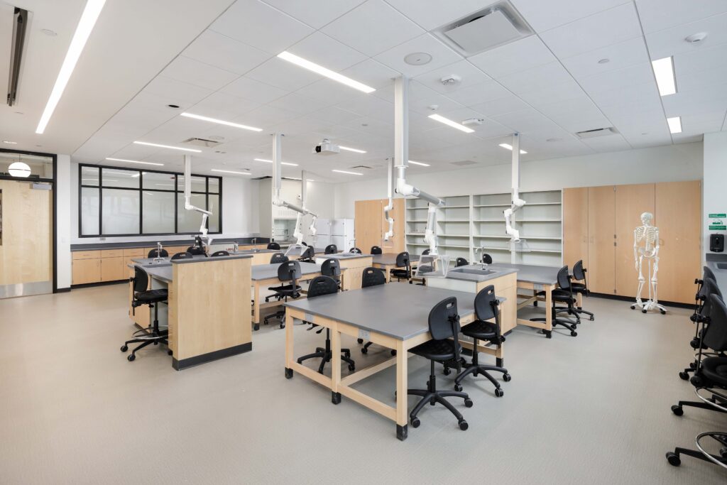 A biology laboratory classroom with multiple workstations, rolling chairs, overhead ventilation snorkels, and a human skeleton model.