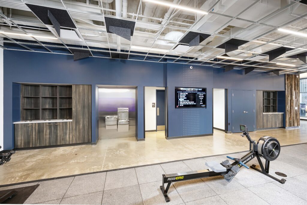 Gym interior with cabinets, a water fountain, a digital “Workout of the Day” screen, and a rowing machine on a concrete floor.