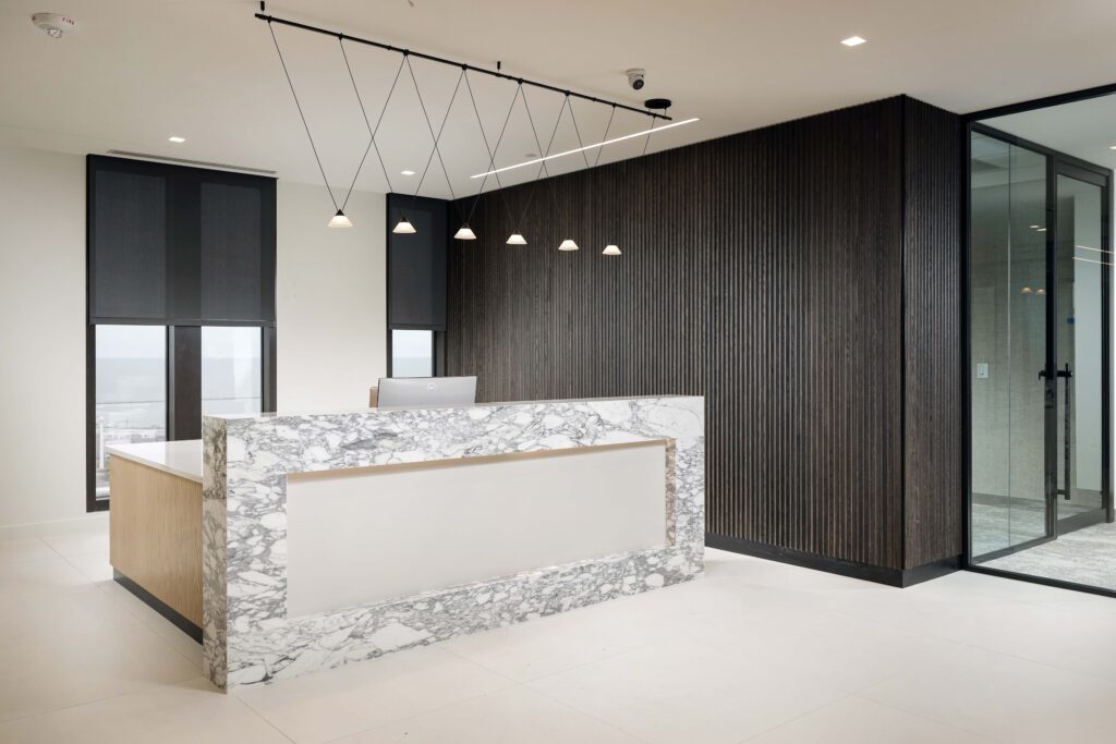 A marble-fronted reception desk in a modern office lobby with dark wood-slatted walls and minimalist pendant lighting.