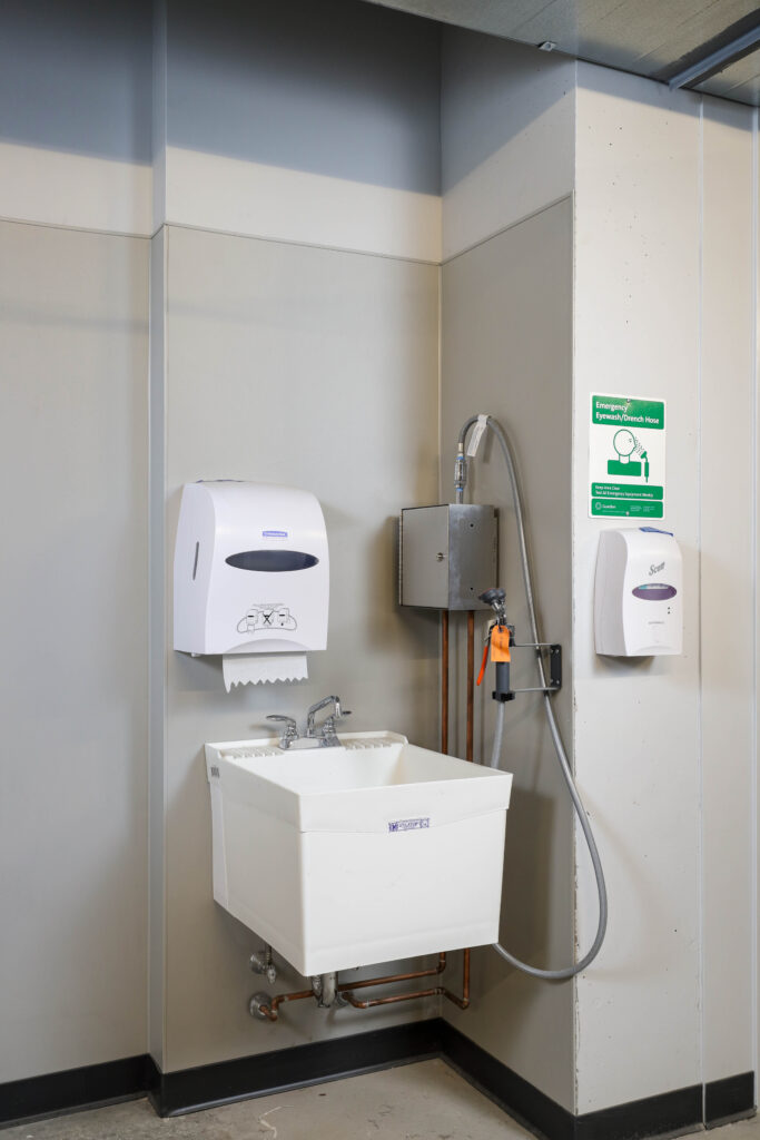 Wall-mounted utility sink with a paper towel dispenser, emergency eyewash drench hose, and instructional sign.