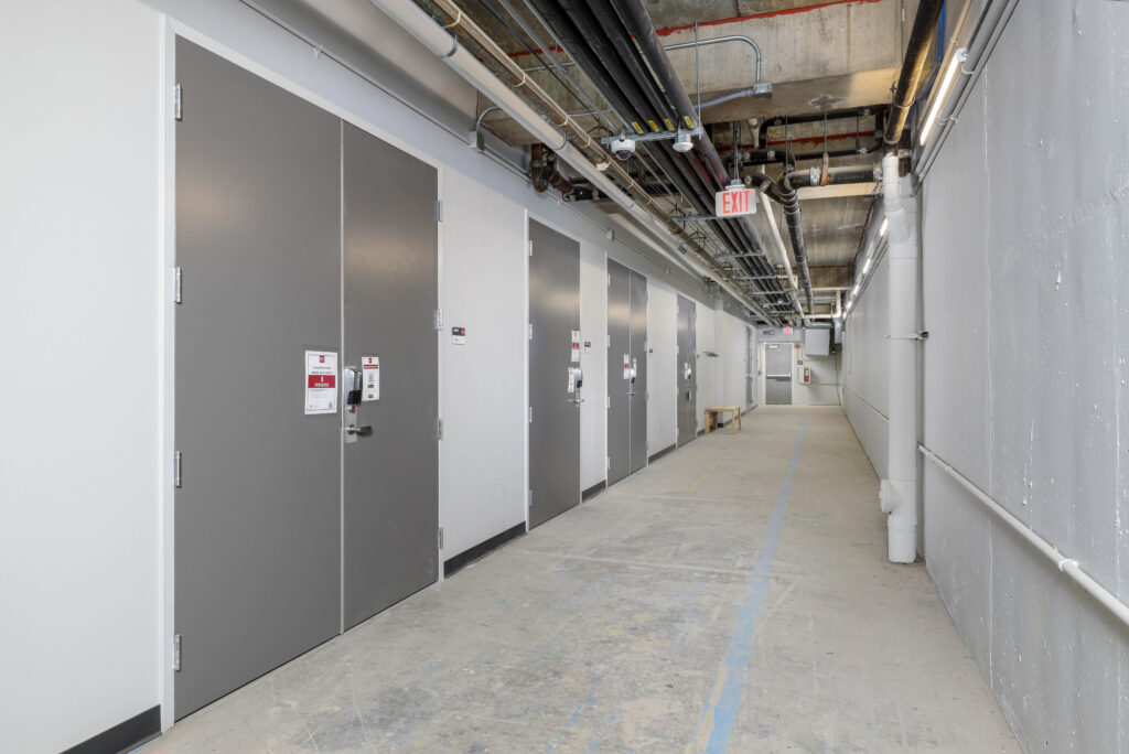 A long, narrow hallway with gray industrial doors along one wall, exposed ceiling pipes, and an illuminated exit sign.