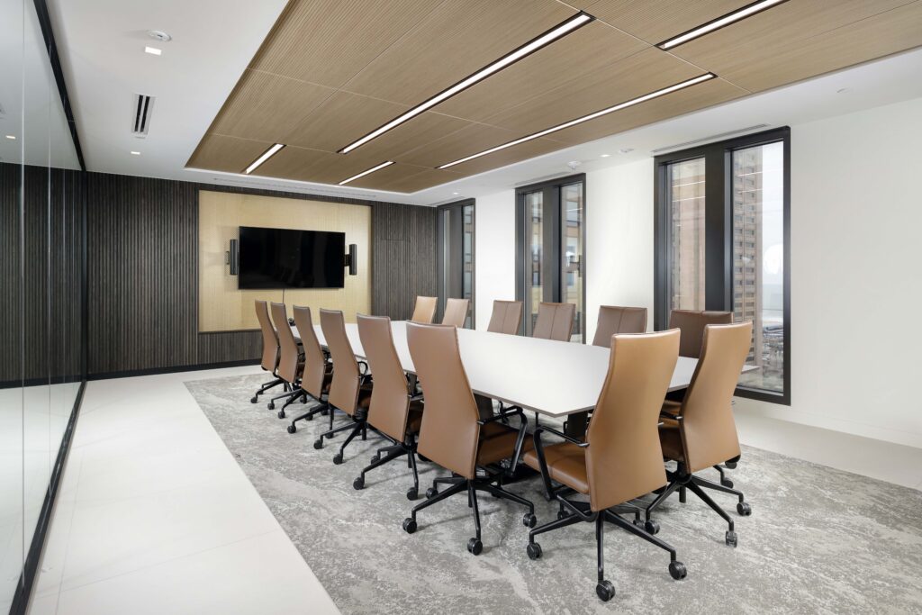 A modern conference room featuring a long white table with tan leather rolling chairs, a wall-mounted television, and floor-to-ceiling windows.