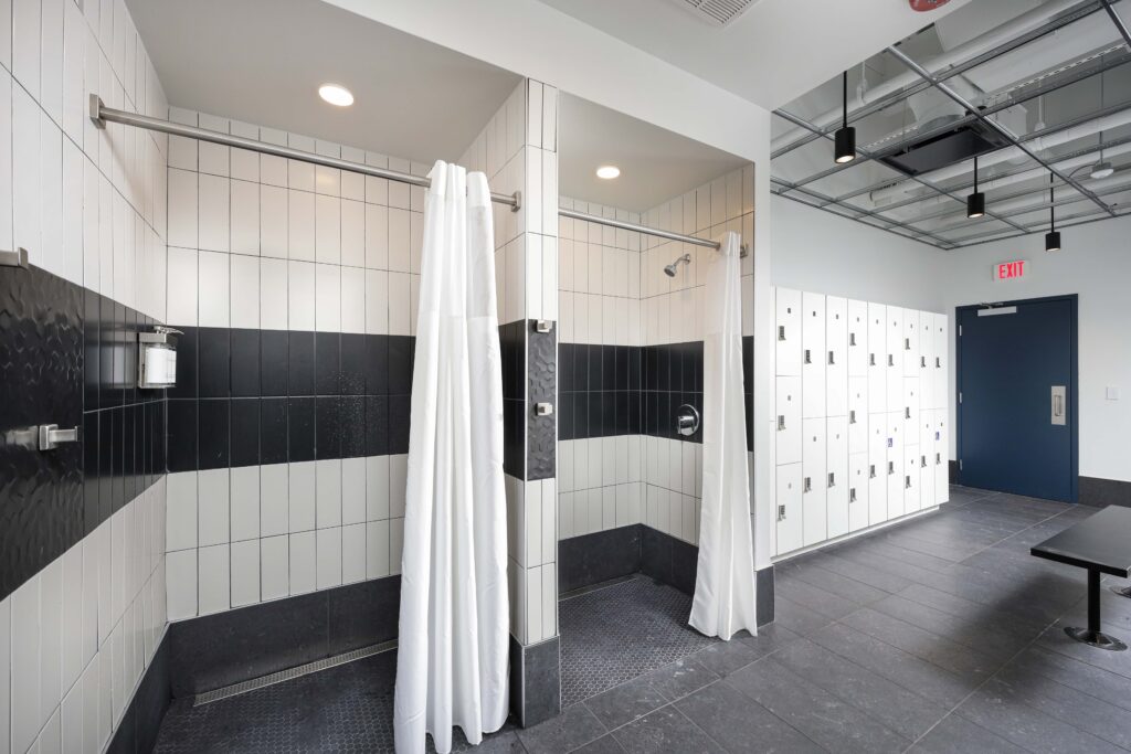 Two shower stalls with white curtains and black and white tiled walls, adjacent to a wall of white lockers and a blue exit door.