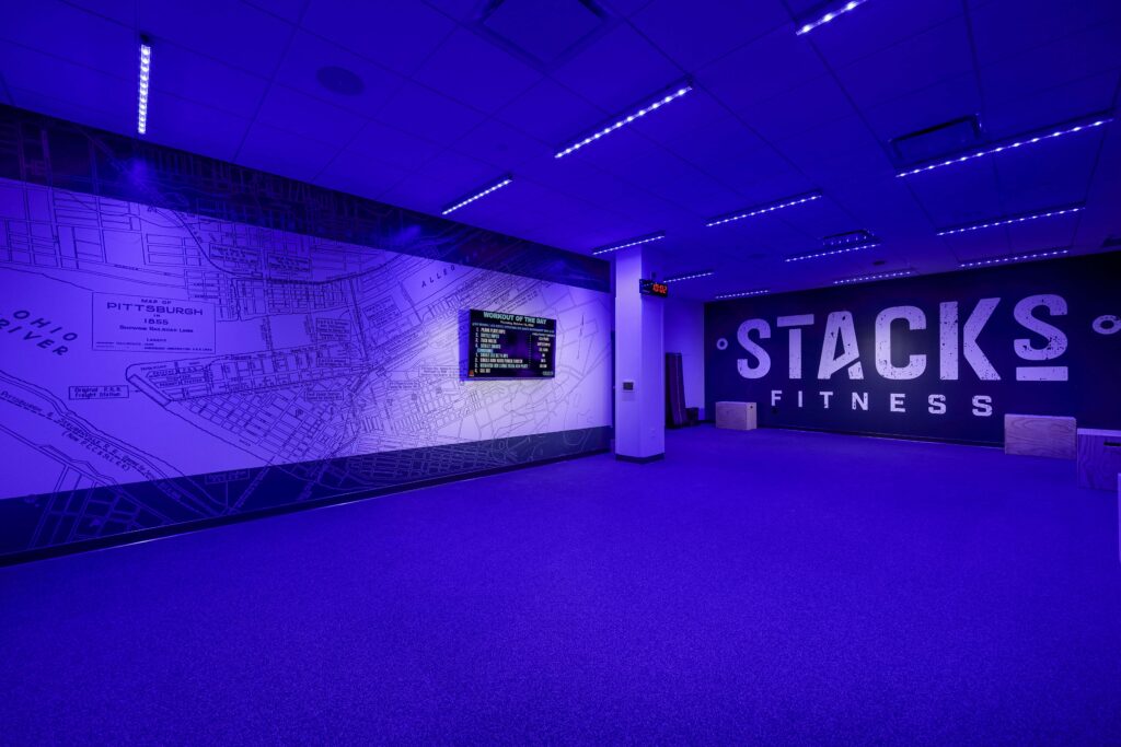 Interior of a gym with purple lighting, a wall-mounted map of Pittsburgh, a “Workout of the Day” digital display, and a wall sign that reads “Stacks Fitness.”