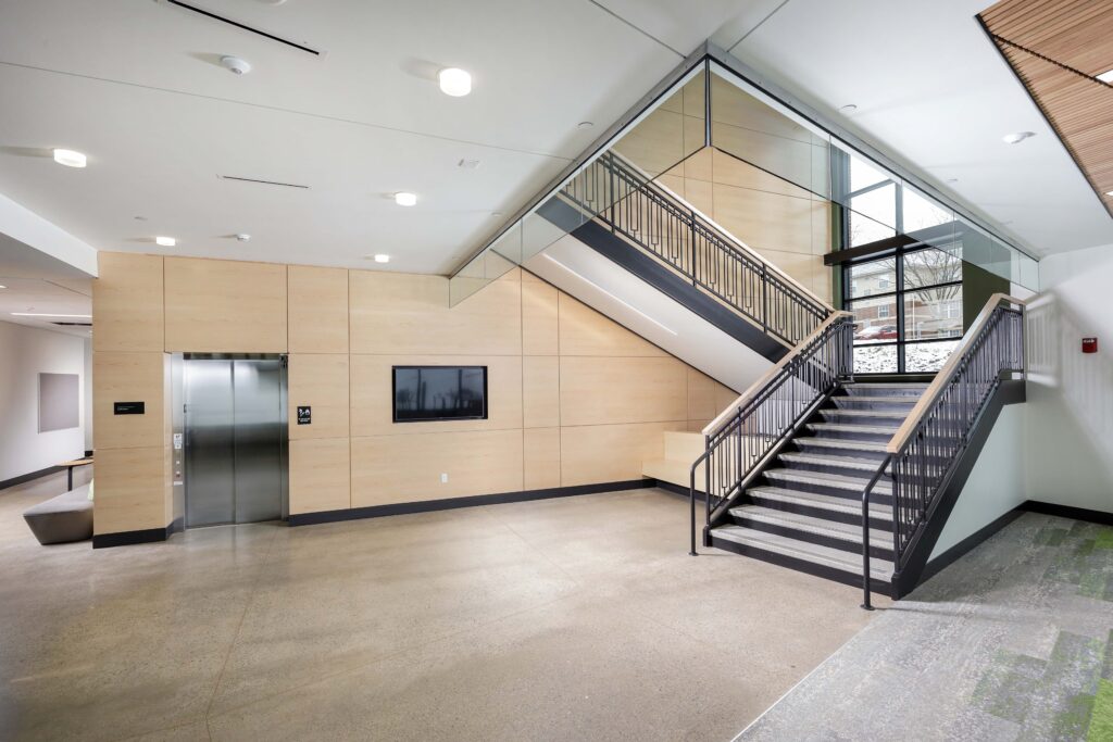 Modern building lobby featuring a wood-paneled wall with an elevator and mounted screen, alongside a metal staircase with railings.