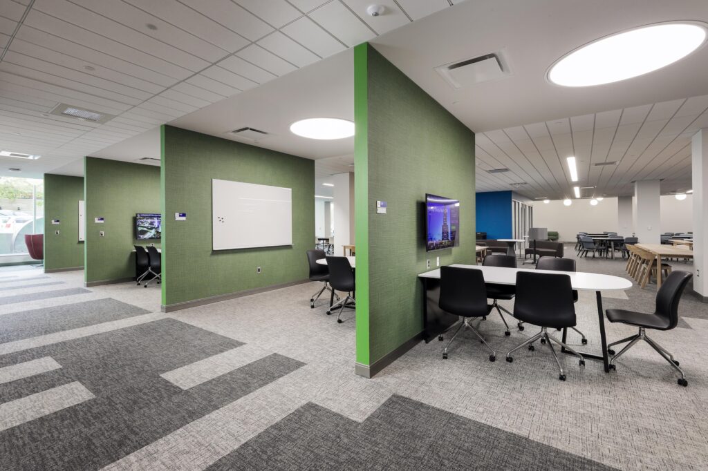 Office interior with green partition walls, whiteboards, computer monitors, and chairs arranged at tables in open workspaces.
