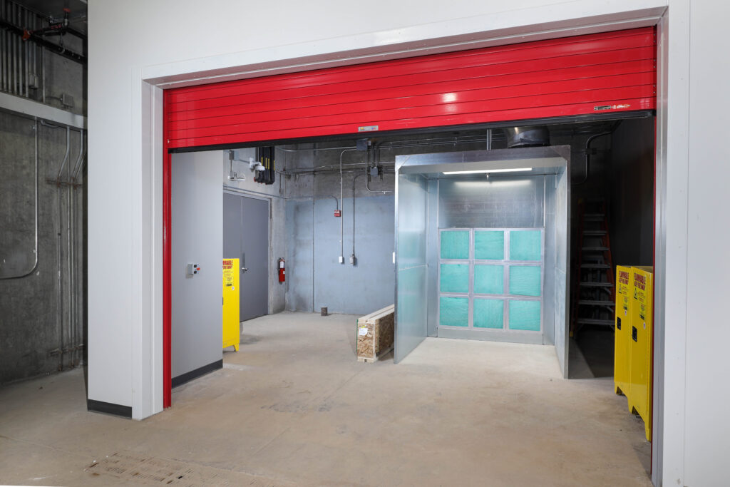 A metal spray booth with green filters sits inside a room with a partially raised red roll-up door.