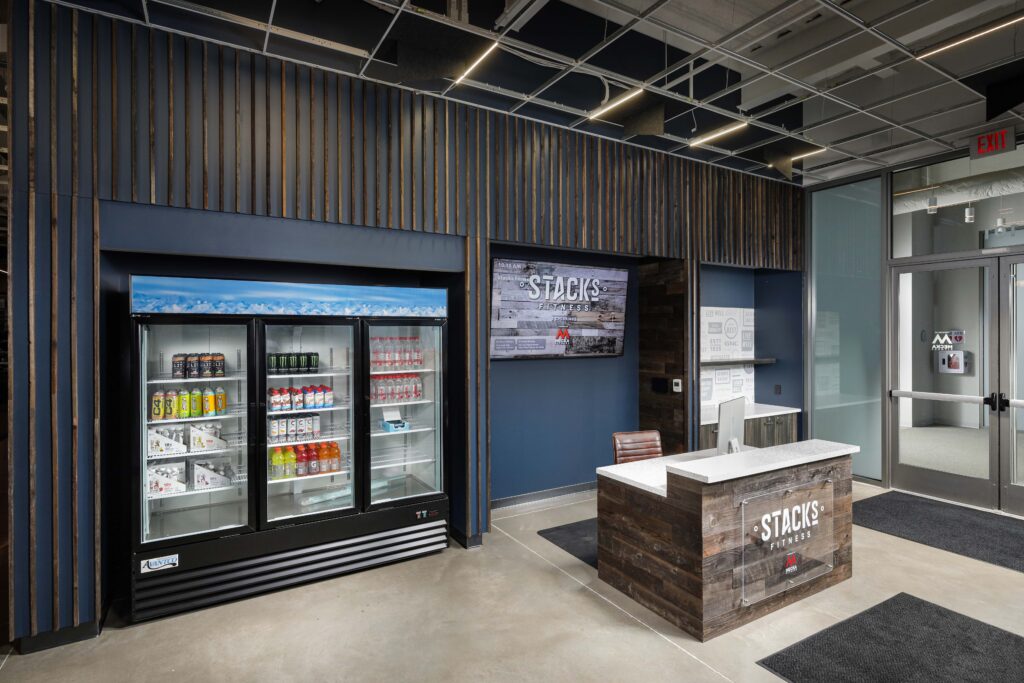 A Stacks Fitness reception area with a glass-front beverage cooler, a wood-paneled front desk, and a wall-mounted digital sign.