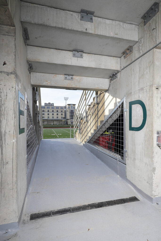 A concrete stadium tunnel entrance marked with the letters E and D, leading to a view of a football field and a multi-story building.
