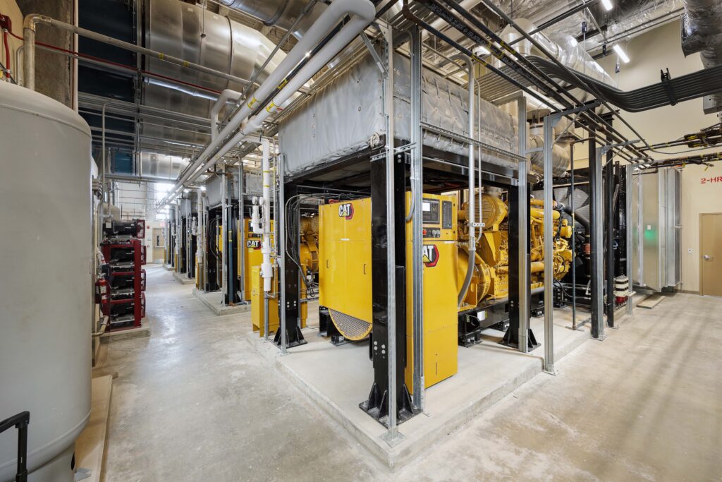Row of industrial Caterpillar diesel generators inside a utility room with exposed overhead pipes.
