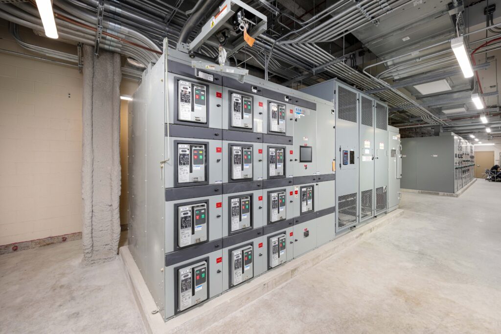 Large industrial electrical switchgear cabinet with multiple circuit breakers, located in a utility room with exposed conduit on the ceiling.