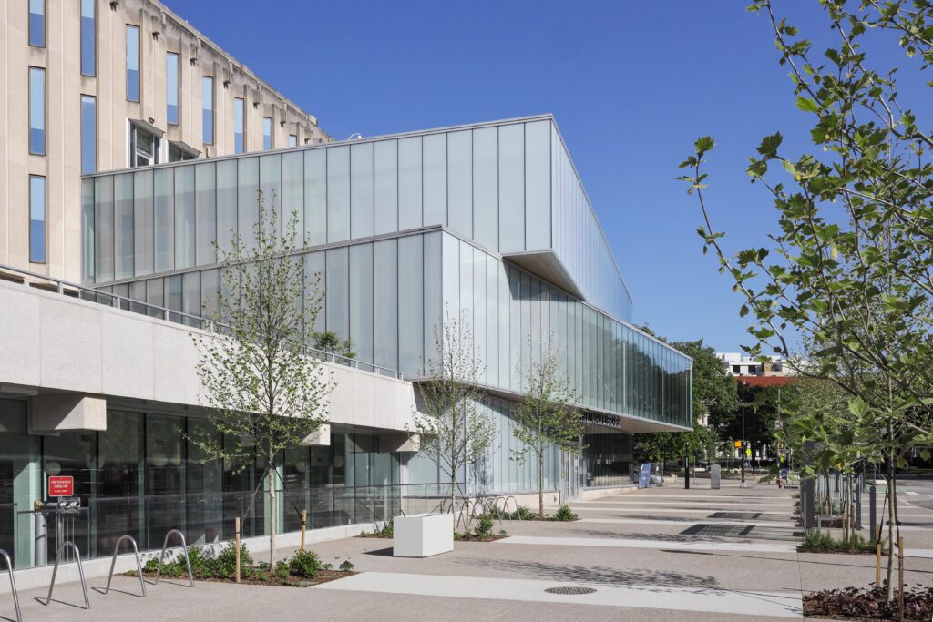 A modern building with a glass facade, glass-enclosed upper levels, and a surrounding paved plaza with young trees.