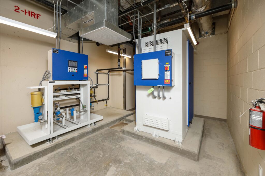 Two industrial fuel pump systems housed in a utility room with exposed piping and a wall-mounted fire extinguisher.