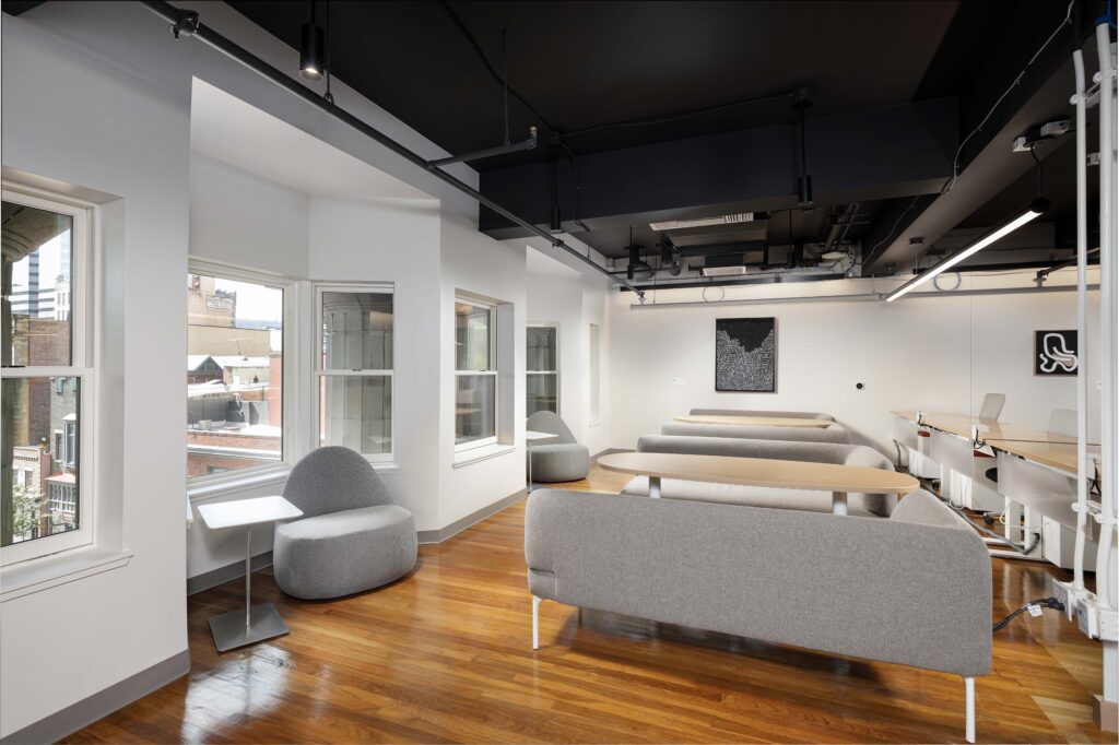 Modern office interior with wooden floors, grey modular seating, and a long desk with white chairs.